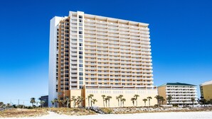 Un grand hôtel à plusieurs étages, doté d’un toit plat et de balcons à chaque étage, situé près d’une plage de sable bordée de palmiers.