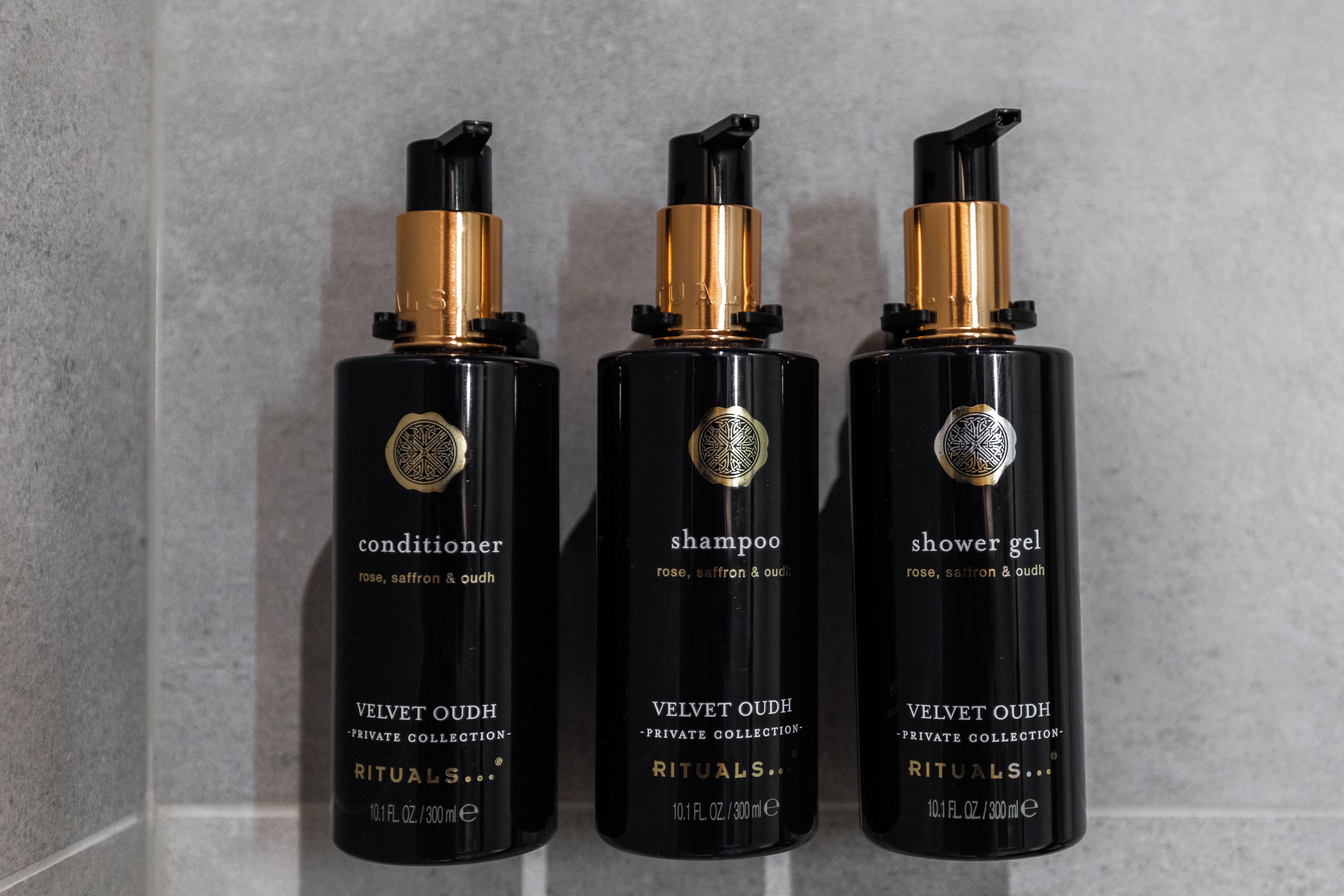 Trois flacons noirs ornés de détails dorés, étiquetés « après-shampoing », « shampoing » et « gel douche », chacun étant doté d’un bouchon doré et d’un logo circulaire.