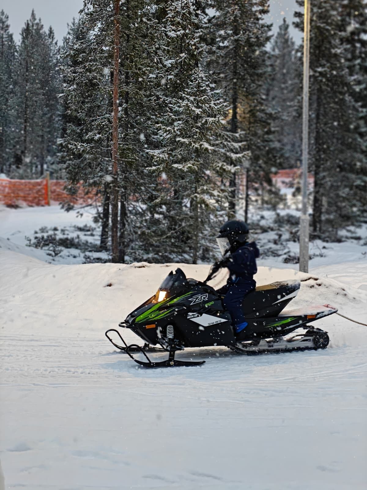 A person riding a snowmobile on a snowy path with trees and an orange fence in the background.