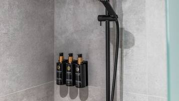 Zona de ducha con cabezal de ducha manual, grifo empotrado y rociador manual. Hay tres botellas de champú y acondicionador en la pared.