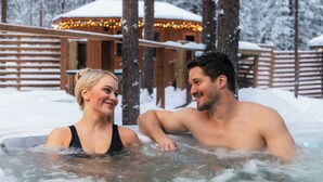 Ein Paar in einer Badewanne, umgeben von schneebedeckten Bäumen und einer Holzhütte mit Lichterketten.