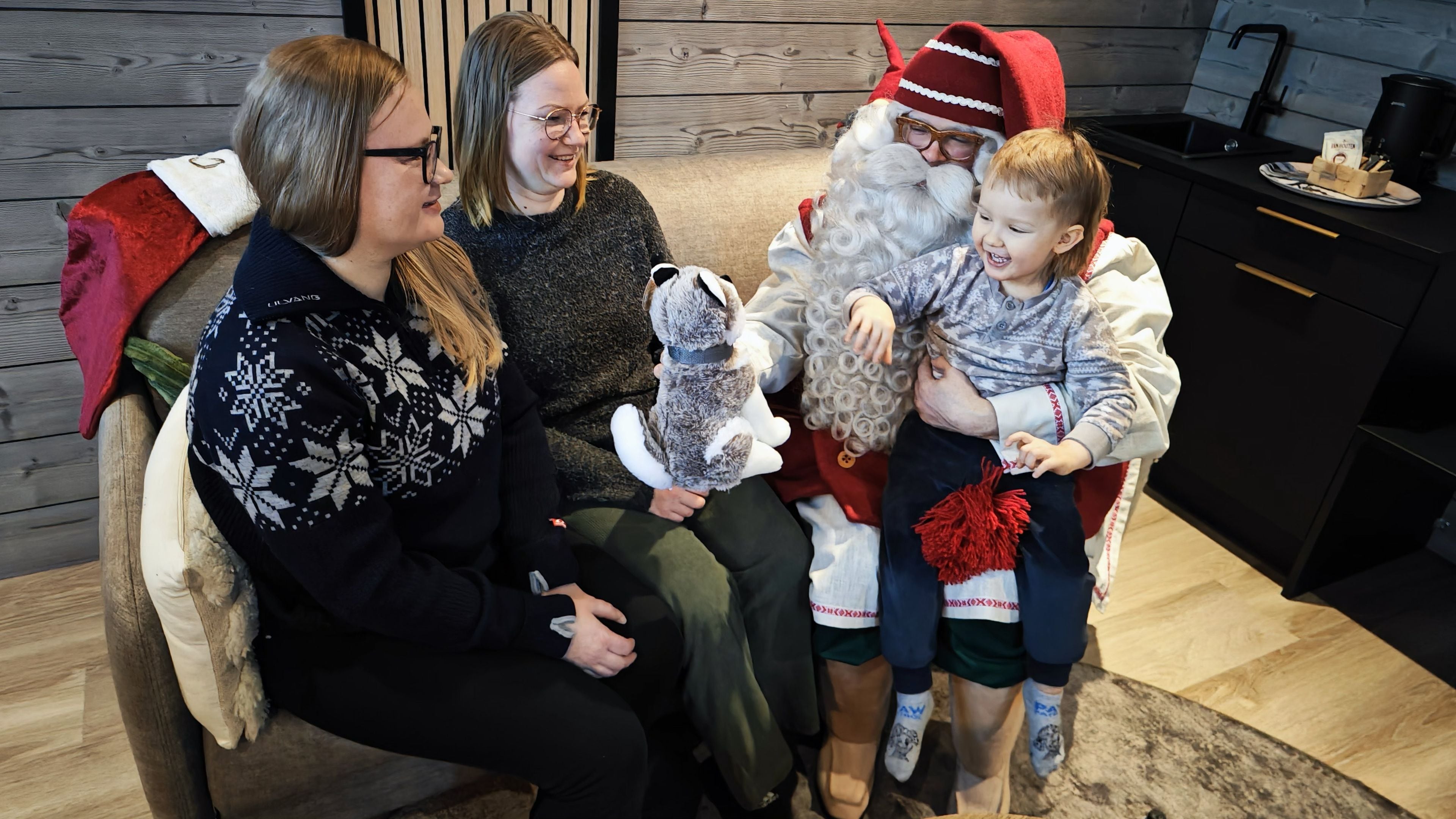 Perhe, jossa joulupukki-hahmo istuu sohvalla, lapsi pitää sylissään pehmolelua ja keittiönurkkauksessa on kahvinkeitin ja keittiövälineitä.