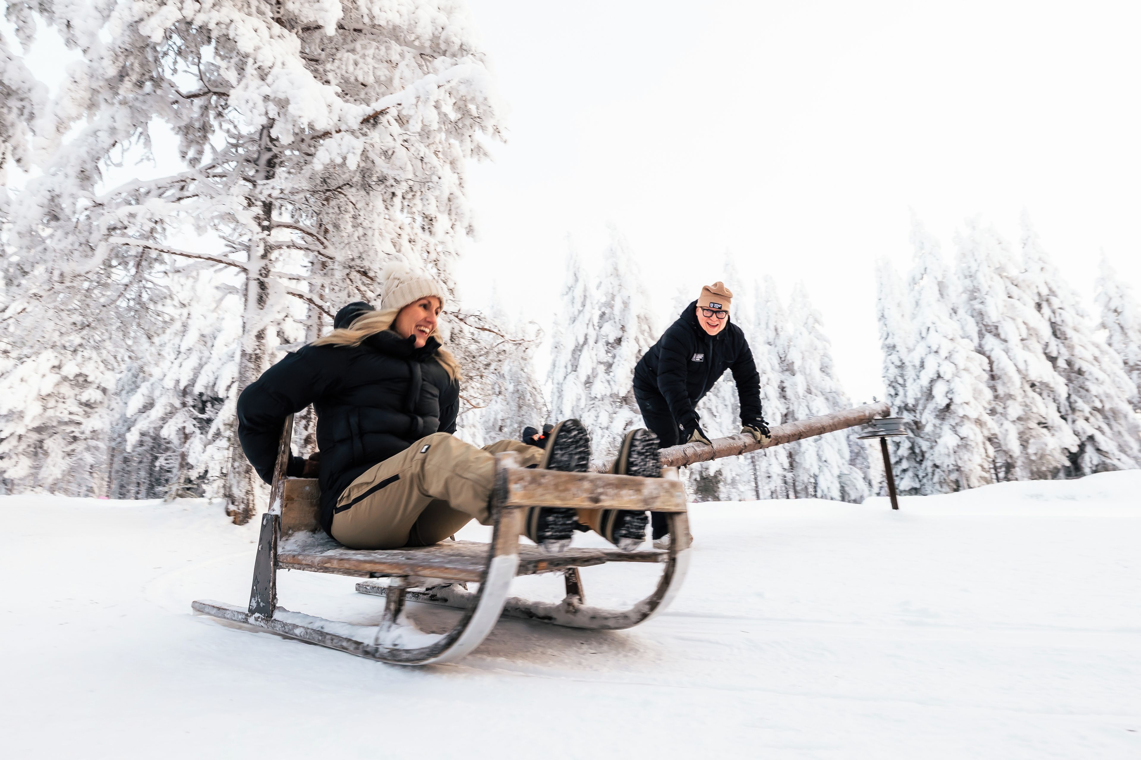Two people on a wooden sled in a snowy landscape with trees.
