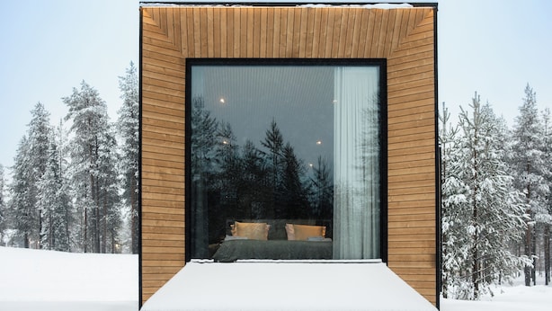 Ein modernes Blockhaus mit großem Fenster, einem Bett und Blick auf schneebedeckte Bäume.