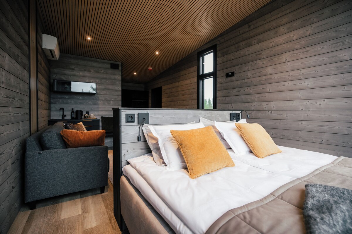 A modern bedroom with a wooden ceiling, a bed with white and mustard pillows, a grey sofa, and a TV mounted on the wall.