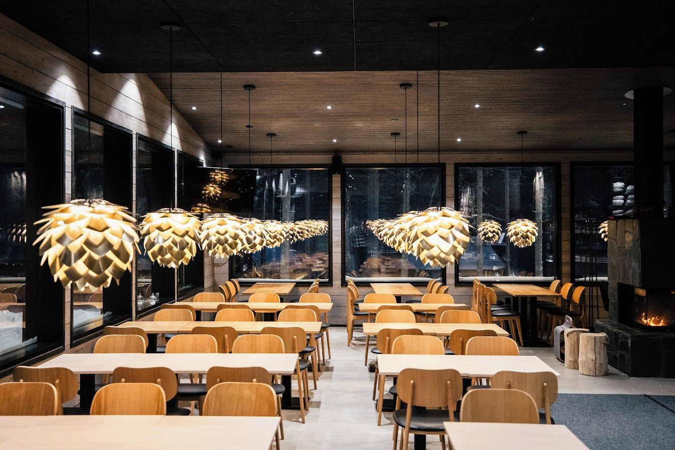 Un espace de restauration moderne avec des tables et des chaises en bois, de grandes fenêtres et des luminaires suspendus au design original.