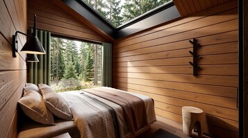 Un dormitorio con techo inclinado, paredes de madera, cama con almohadas, lámpara de noche y ventana con vista a árboles.