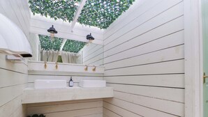 Un baño pequeño con paneles blancos, dos lavamanos, un espejo y un techo decorado con follaje artificial.