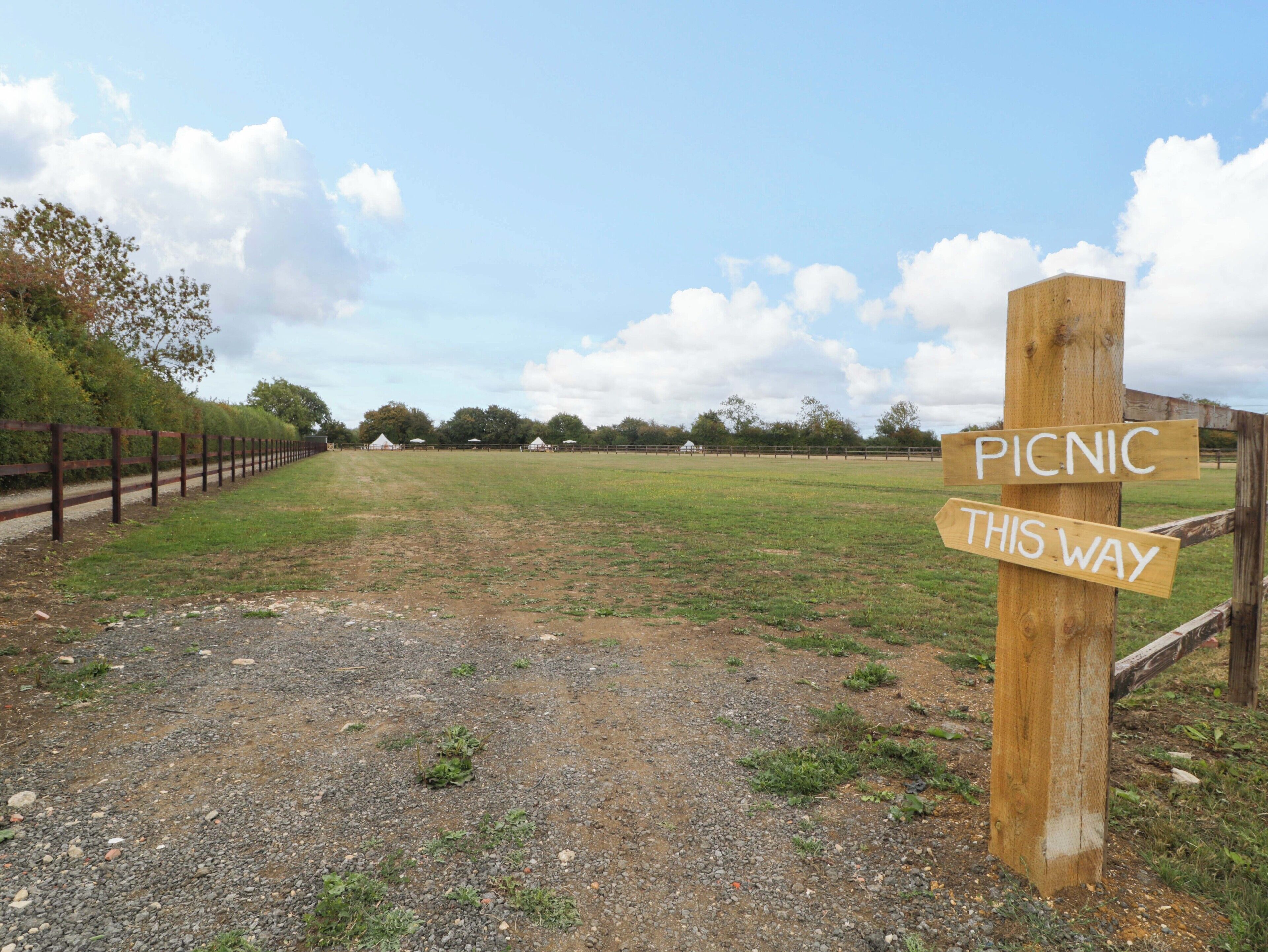 A rural landscape with a dirt path leading to a picnic area, surrounded by a wooden fence and trees.