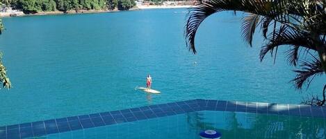 Piscina azul cristalina con una persona haciendo paddle surf, rodeada de exuberante vegetación y montañas al fondo.