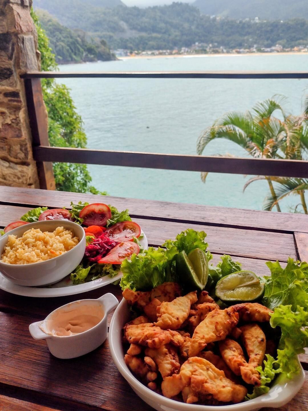 A table with a meal consisting of rice, salad, and fried chicken, set against a scenic view of a beach and mountains.