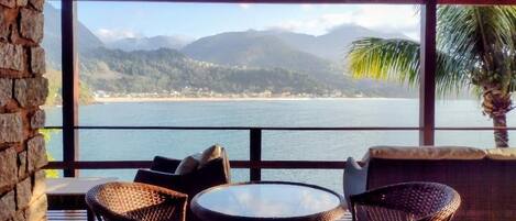 Une terrasse en bois, agrémentée de chaises en osier et d’une table ronde, offrant une vue imprenable sur la côte, avec des montagnes et une plage.