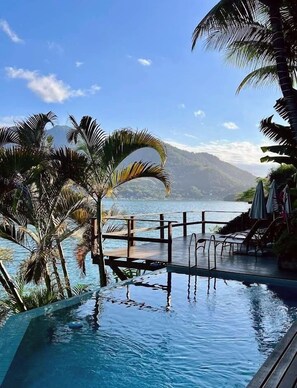 Piscina con cubierta de madera y sillas de descanso, rodeada de palmeras y vistas a una montaña y agua.