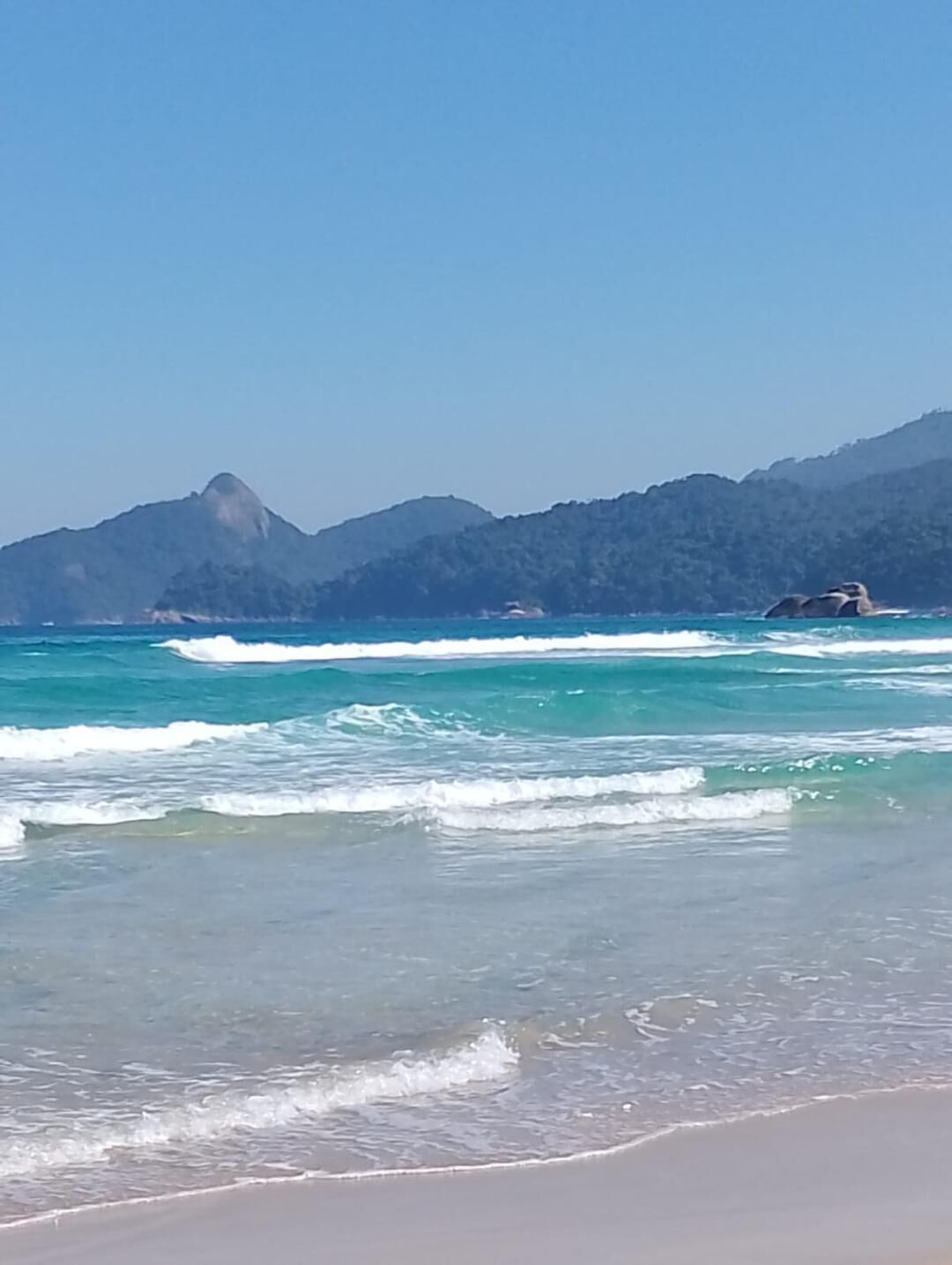 Una playa con aguas turquesas cristalinas, olas suaves y un telón de fondo montañoso.
