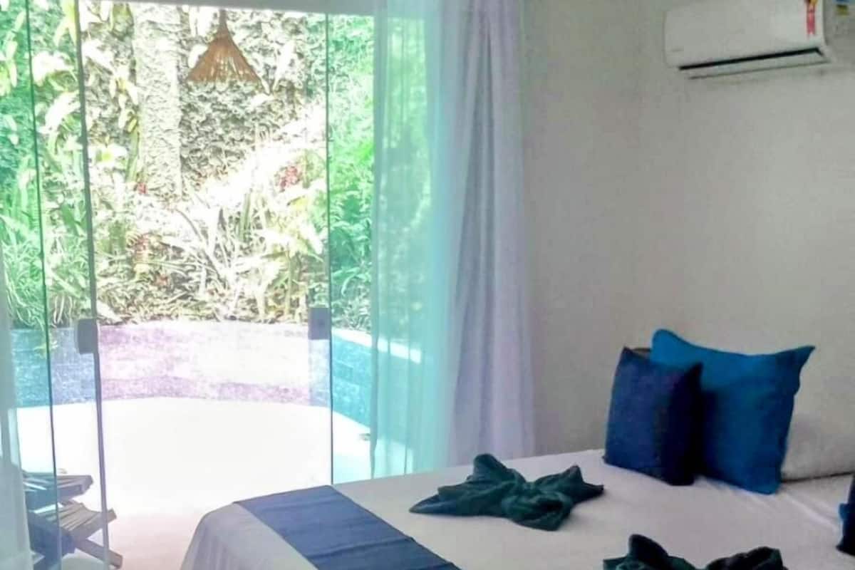 A bedroom with a bed, a hanging light fixture, a sliding glass door leading to a balcony, and an air conditioning unit on the wall.