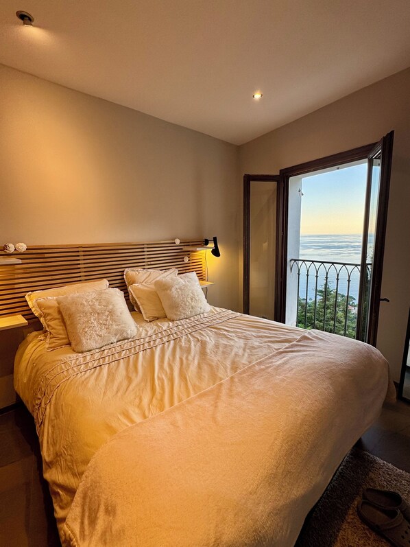 A bedroom with a large bed, a nightstand, a lamp, and a view of the sea through a balcony.