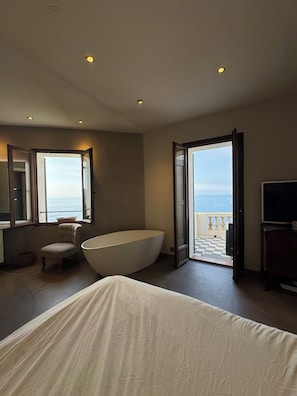 A bedroom with a large bed, a bathtub, a chair, a television, and a balcony with a view of the sea.