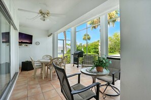A sunlit patio with a ceiling fan, a table with chairs, a grill, and a view of greenery.