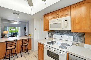 A modern kitchen with wooden cabinets, a white microwave, a stove, and two bar stools. - Naples Retreat - Walk to Pool and More (Naples)