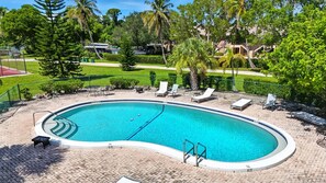 Une piscine avec des chaises longues et une terrasse dallée, entourée d’arbres et avec un bâtiment en arrière-plan.