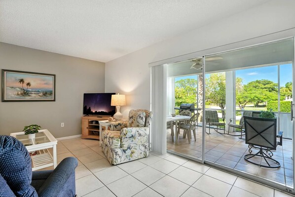 A living room with a TV, a sofa, a chair, a table with a plant, and a balcony with outdoor furniture. - Naples Retreat - Walk to Pool and More (Naples)