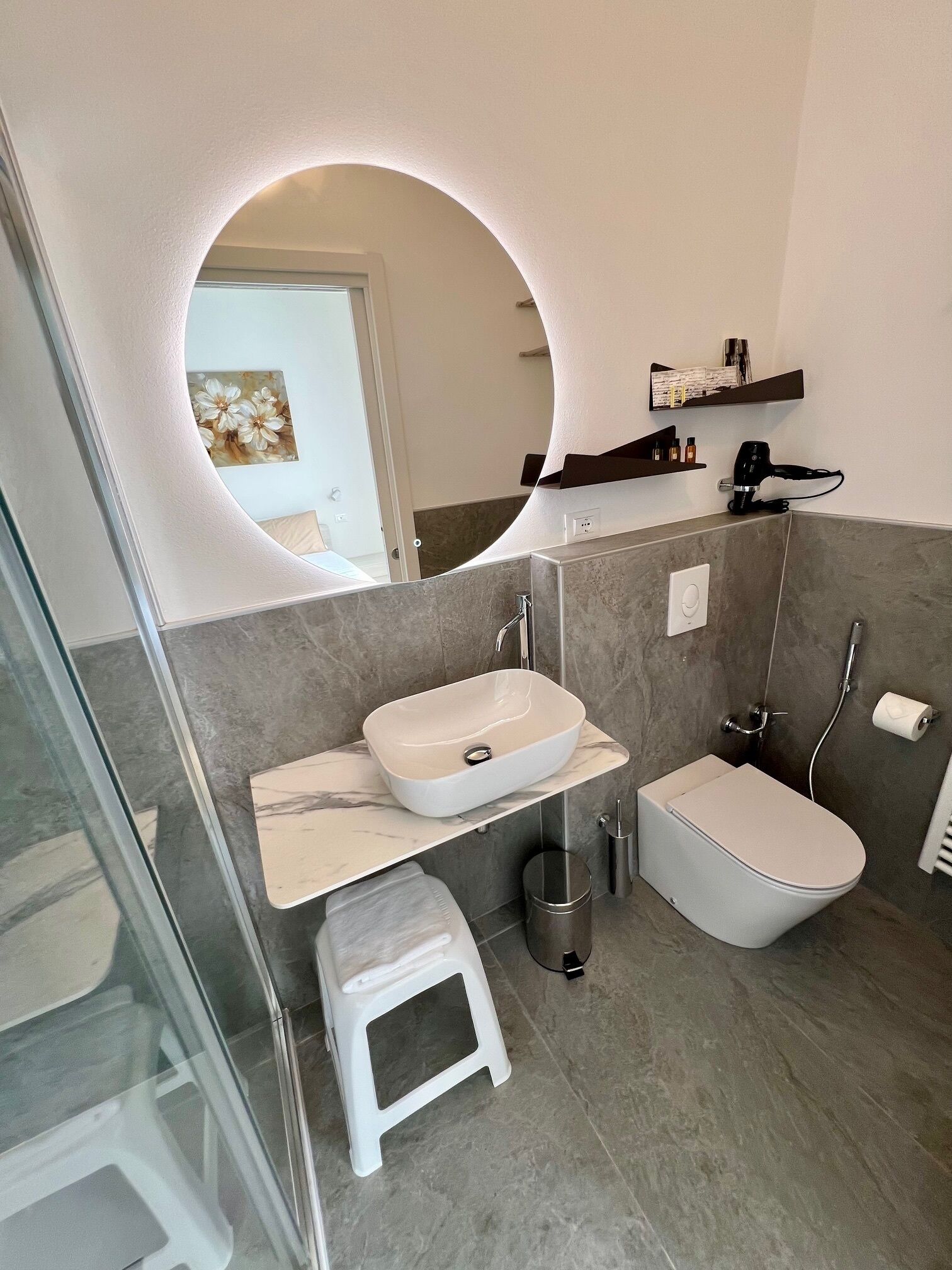 A modern bathroom with a white sink, a toilet, and a shower. There is a small stool and a trash can.