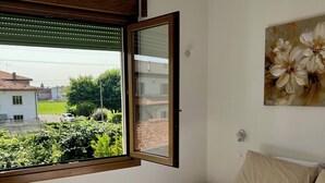 Une chambre avec un grand lit, une fenêtre avec des stores, un tableau de fleurs et une vue sur une maison et de la verdure à l’extérieur.