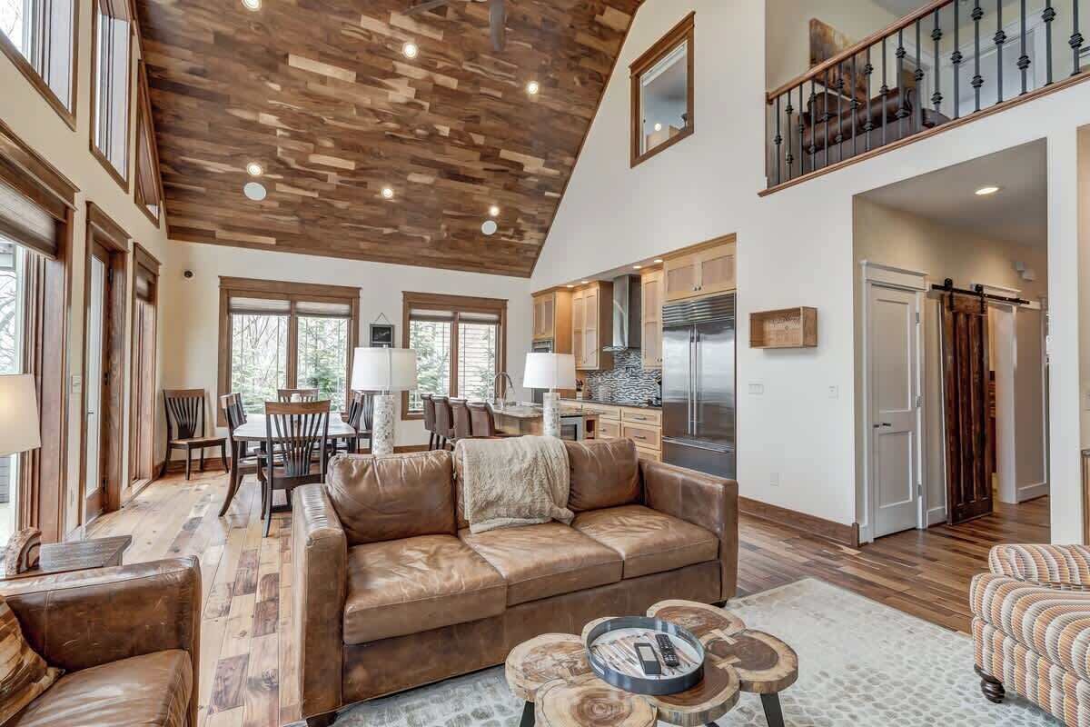 A spacious living area with a high ceiling, wooden flooring, a leather sofa, a dining table, and a kitchen in the background.