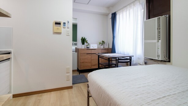 Ein Hotelzimmer mit Bett, Schreibtisch, Stuhl, kleinem Tisch, Heizkörper, Fenster mit Vorhängen, Pflanze und Wandthermostat.