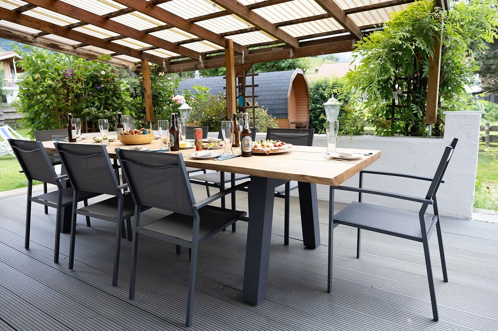Eine Terrassengruppe im Freien mit Holztisch und -stühlen, umgeben von Pflanzen und einer Pergola.