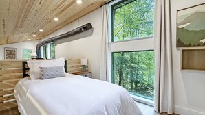 A bedroom with a large bed, wooden beams on the ceiling, a window with a forest view, and a wall-mounted air vent. - Coal Ridge Cabin - WV Retreat with Family History (Summersville)