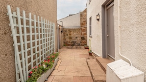 Un estrecho patio con una valla de madera blanca, un muro de piedra, una pequeña mesa con sillas y plantas en macetas.