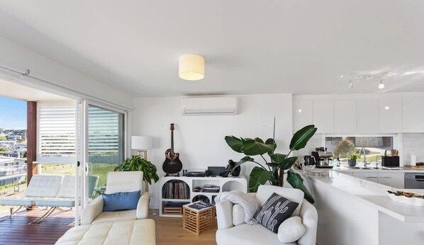 Ein modernes Wohnzimmer mit einer Couch, einem Couchtisch und einem Bücherregal. Eine große Pflanze, eine Gitarre und ein Fenster mit Blick ins Freie sind ebenfalls zu sehen.