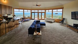 Un amplio salón con un sofá grande, una mesa de centro y un televisor. Varias ventanas ofrecen vistas al exterior.