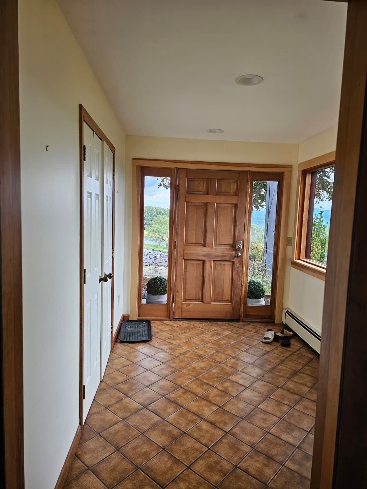 Un pasillo con puerta de madera, suelo de baldosas y vista a follaje exterior.