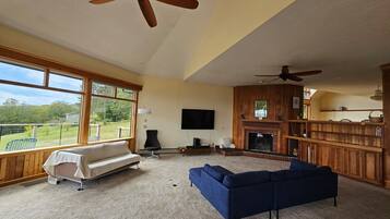 A spacious living room with a large window, a fireplace, a television, and a ceiling fan.