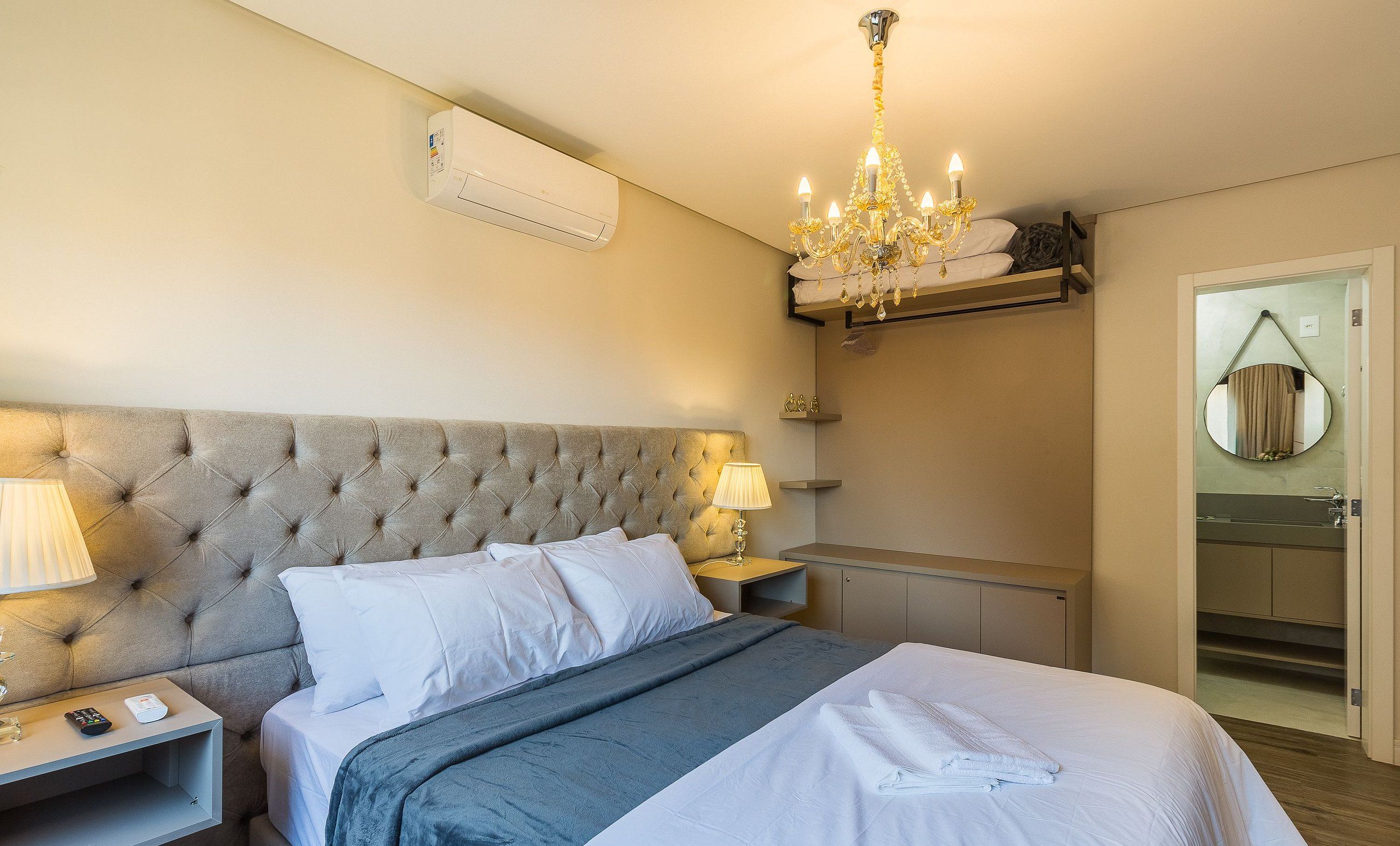 Une chambre à coucher avec une tête de lit capitonnée, une table de chevet avec une lampe, un lustre, un miroir et une salle de bain visible à travers une porte ouverte.