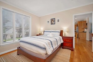 Un dormitorio con cama, mesitas de noche, una ventana con persianas y una cocina visible a través de una puerta abierta, además de una pared decorada con cuadros.