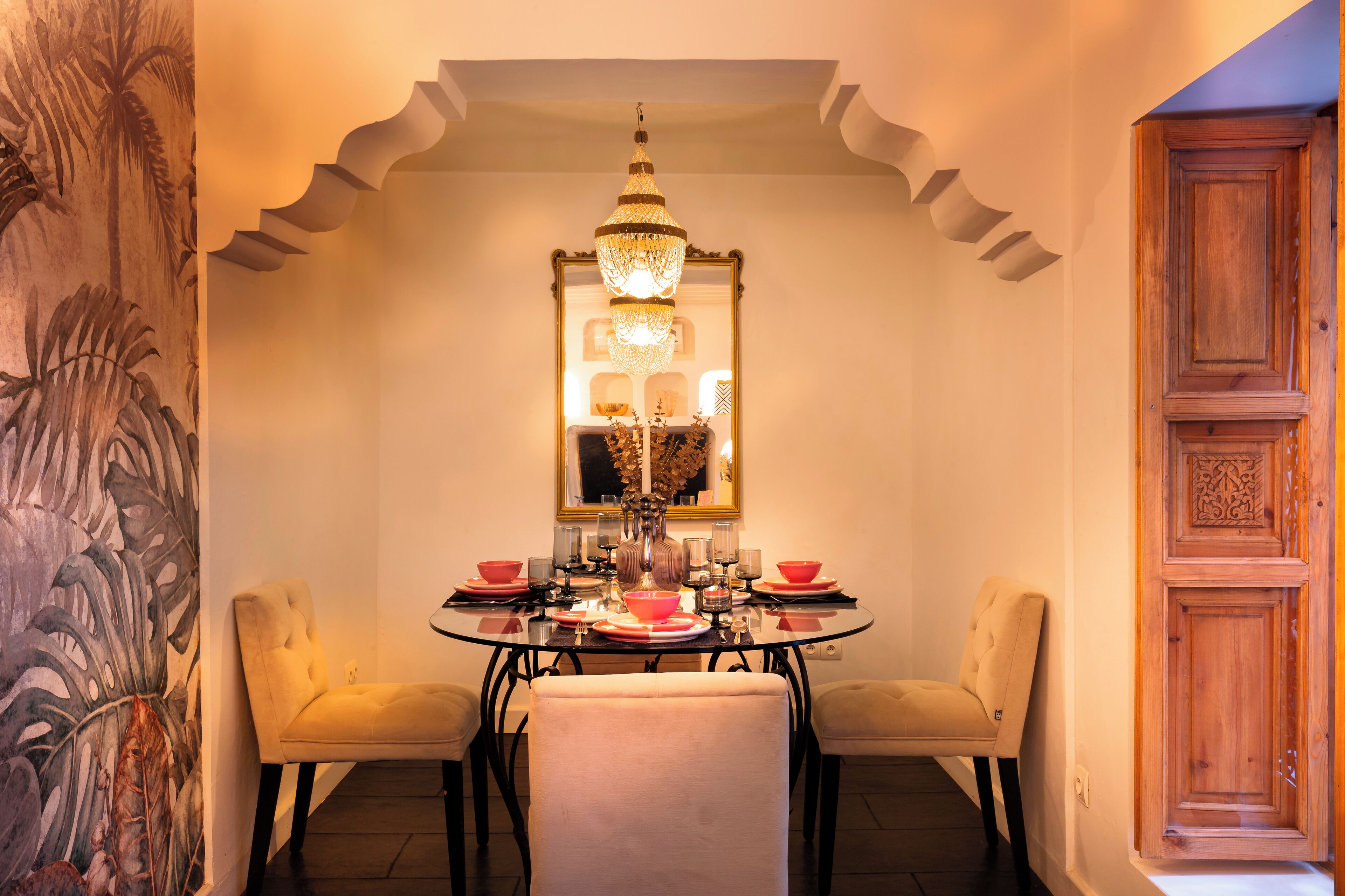 A dining area with a round table, two chairs, a chandelier, and a decorative mirror.