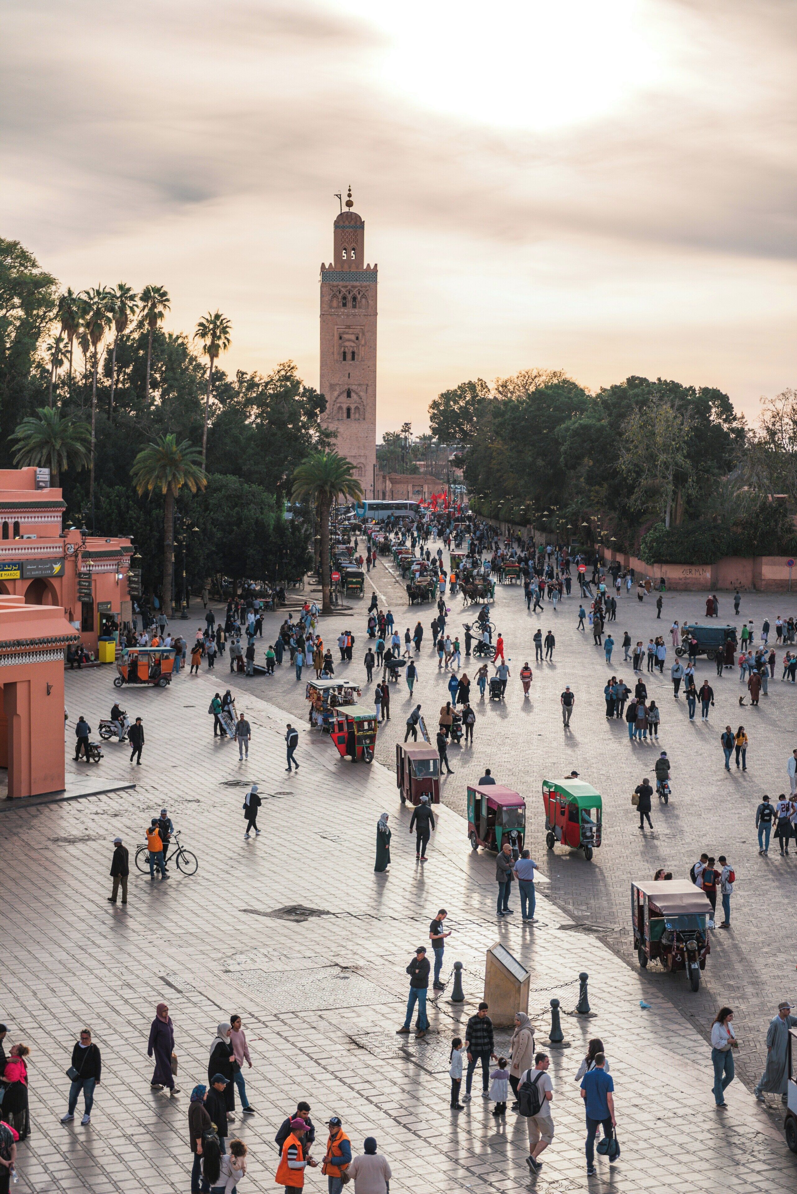 A bustling square with a prominent tower, surrounded by palm trees and various people and vehicles.