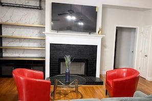 Una sala de estar con chimenea de ladrillo negro, un televisor de pantalla plana montado encima y sillones rojos.