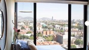 A modern bedroom with a large bed, a small ottoman, and a city view through large windows.