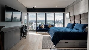 A modern bedroom with a large bed, a desk, and a sofa. It features a city view through large windows.