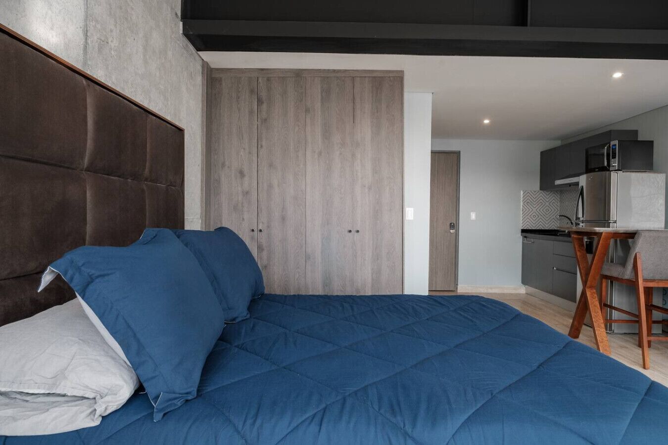 Een moderne slaapkamer met een groot bed, een houten garderobe en een kitchenette op de achtergrond.