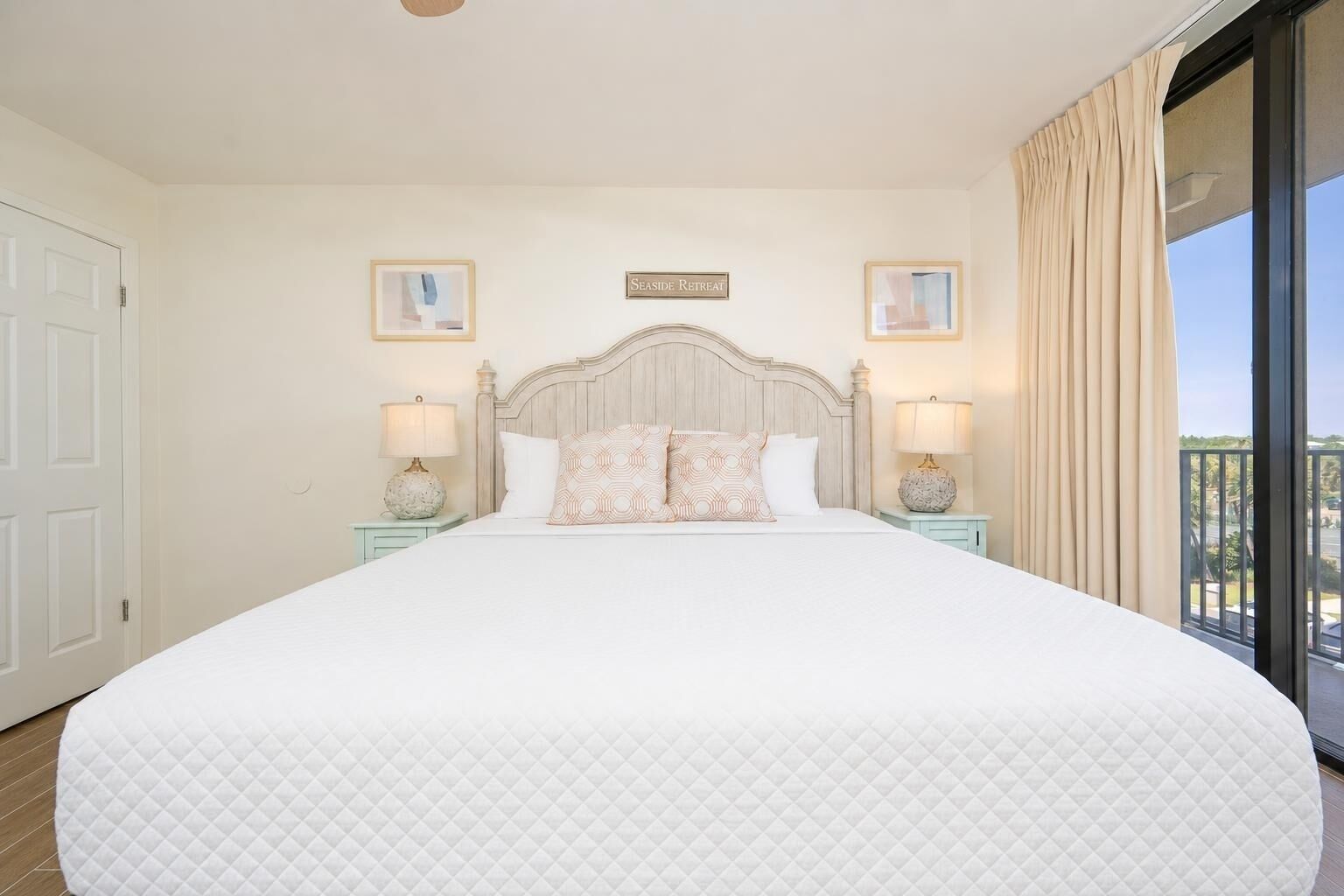 A large bed with a quilted white cover, two bedside tables with lamps, a headboard with a sign, and a balcony with curtains.