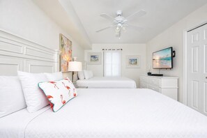 Habitación de hotel con dos camas, ventilador de techo, televisor y obras de arte en las paredes.