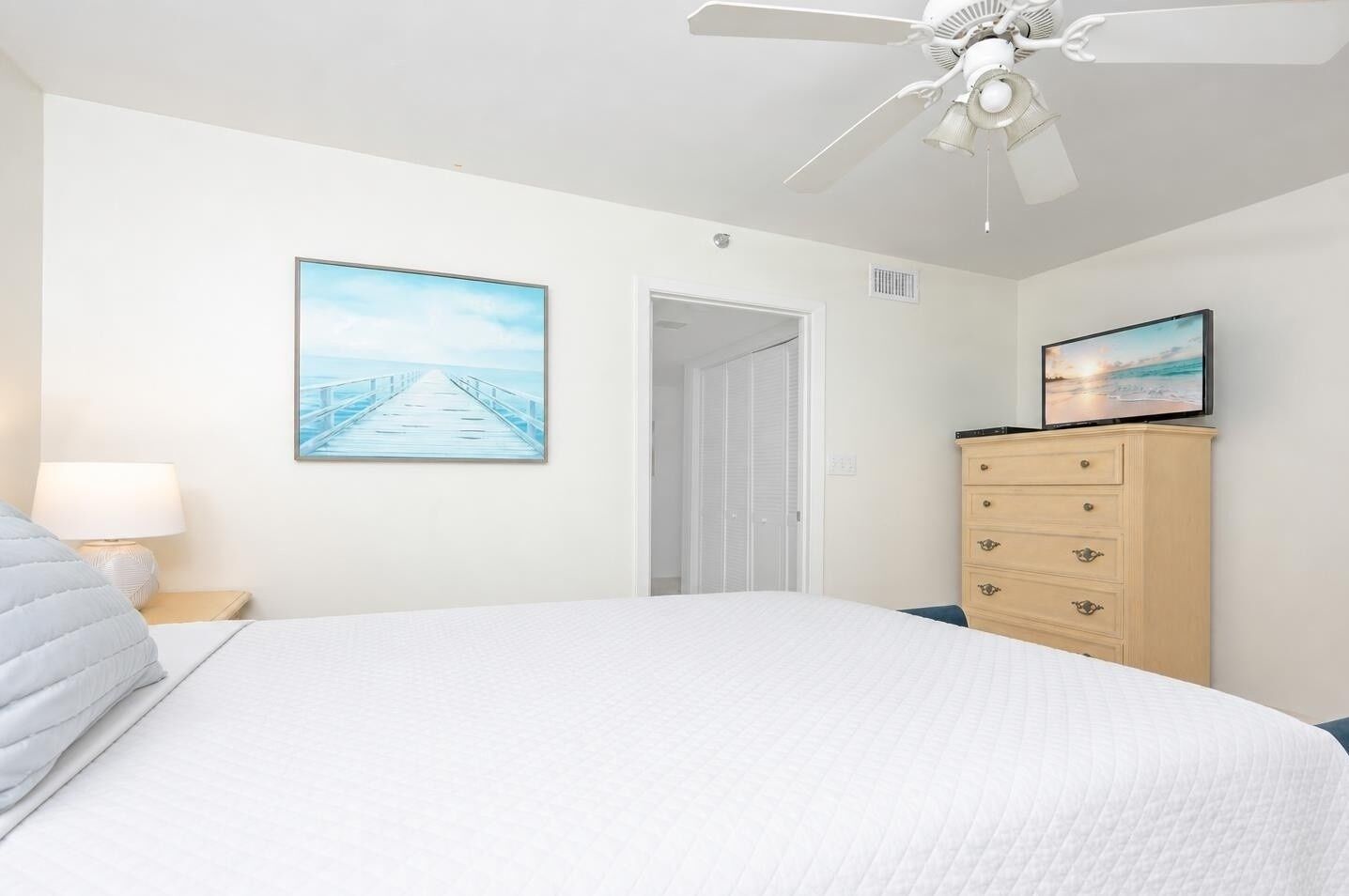 A bedroom with a bed, a dresser, a ceiling fan, a painting, and a door.