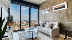 Una sala de estar moderna con un ventanal grande, un sofá, una mesa de centro de vidrio y vistas a un paisaje urbano.