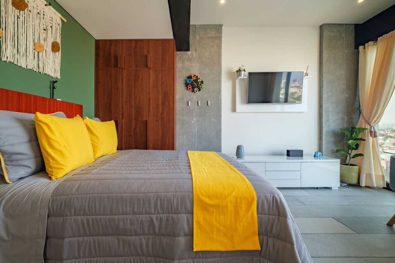 Un dormitorio moderno con una cama grande, un televisor montado en la pared y vistas a la ciudad desde la ventana.