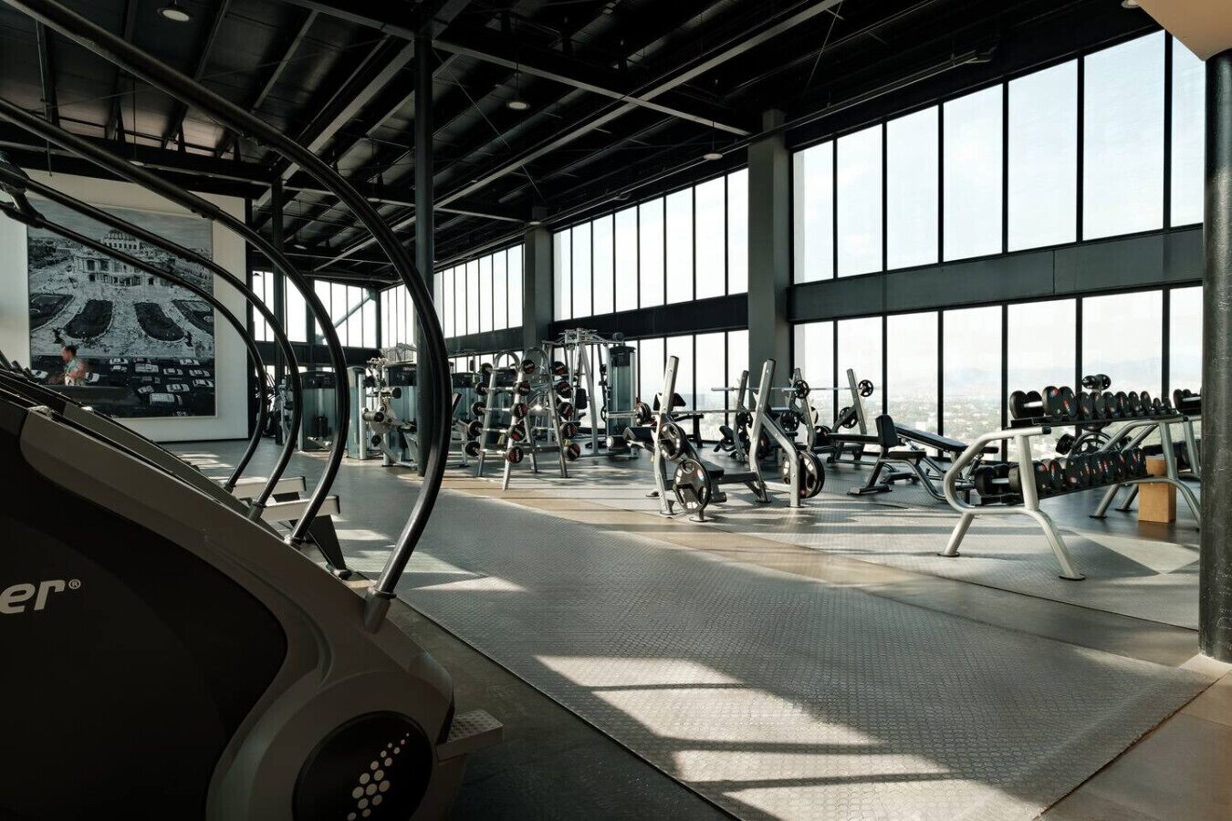 A gym with various exercise equipment, large windows, and a high ceiling.